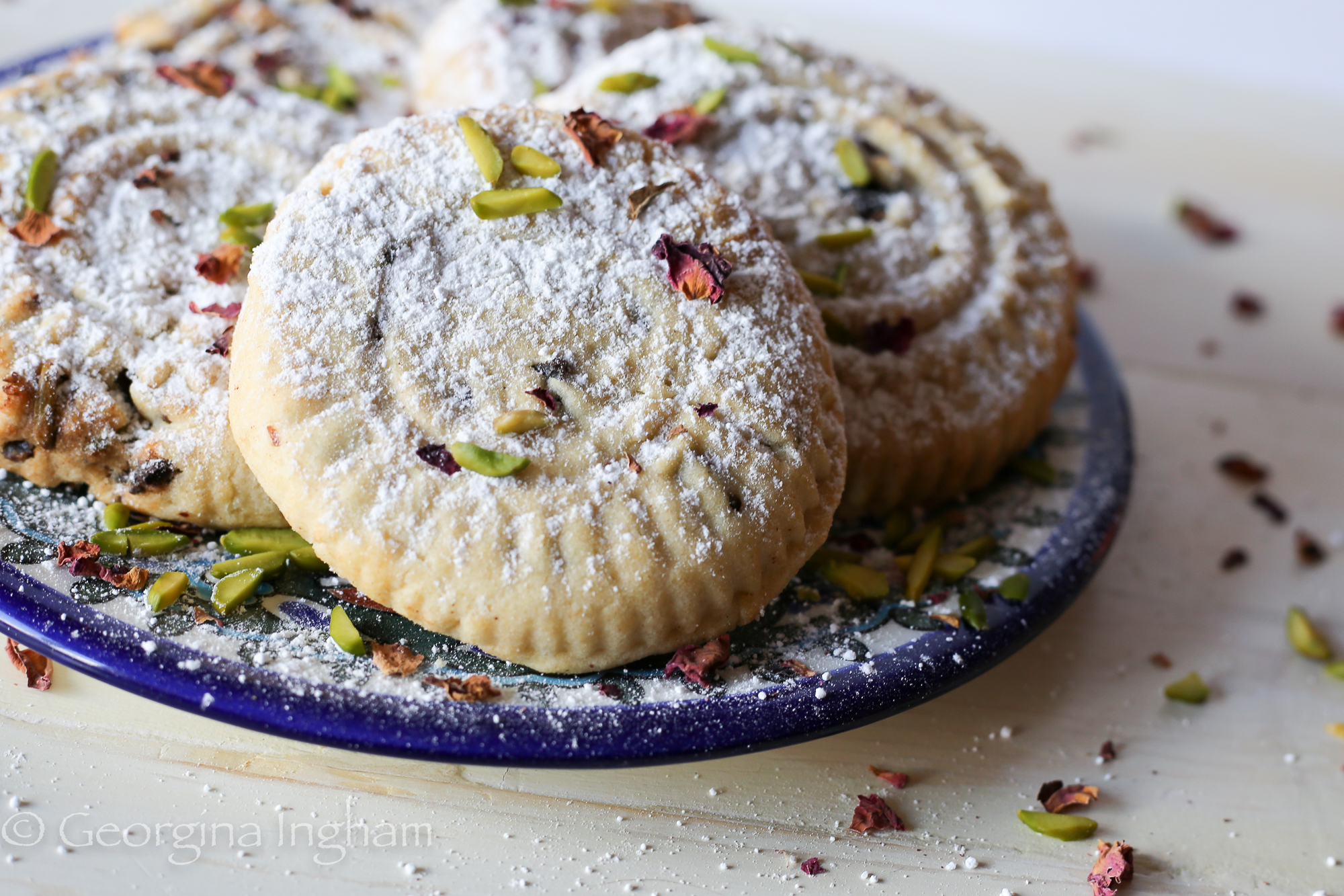 Plate of Ma'amoul cookies scattered with icing sugar, crushed pistachios, and delicate rose petals, styled on a rustic surface